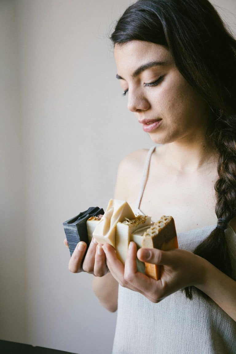 A young woman holding various artisan soap bars, framed indoors with soft lighting.