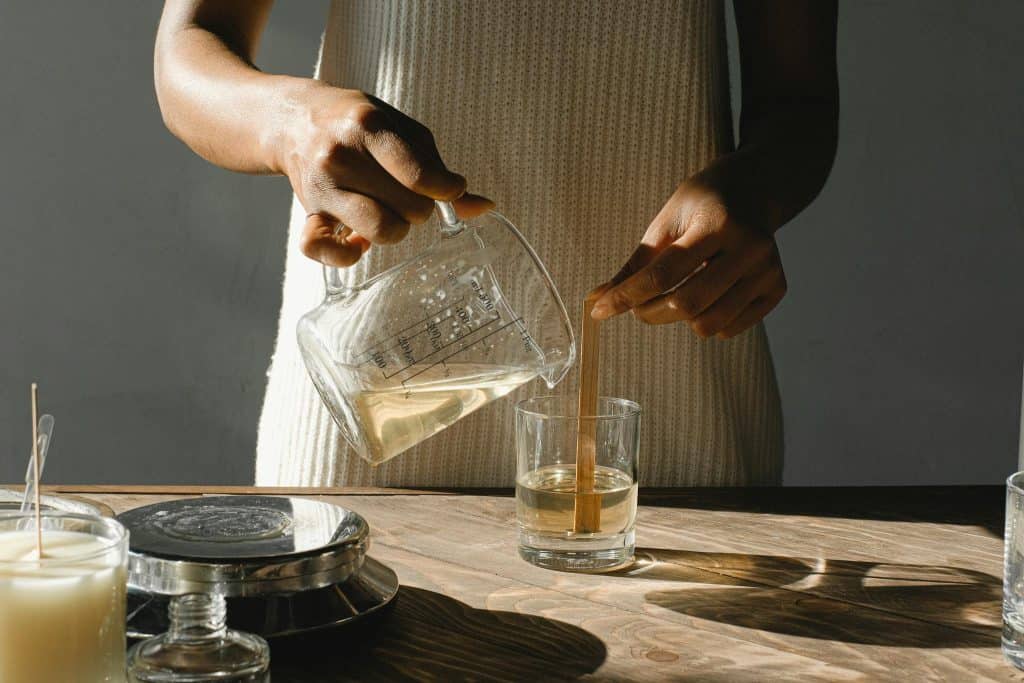 Unrecognizable African American female pouring melted wax from beaker into glass mold with wooden wick while making candles at table